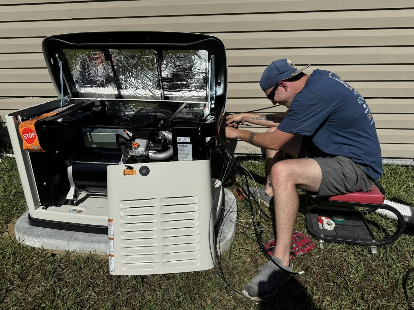 A person in casual clothing works on wiring in an open generator next to a building, seated on a low stool, with grass underneath.