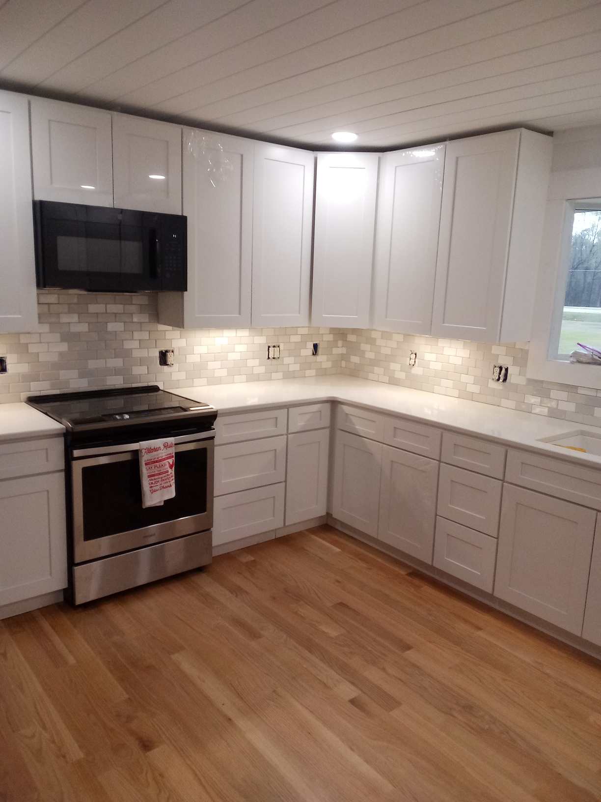 A modern kitchen with white cabinets, stainless steel stove, black microwave, and light wood flooring. The backsplash features white and beige tiles.