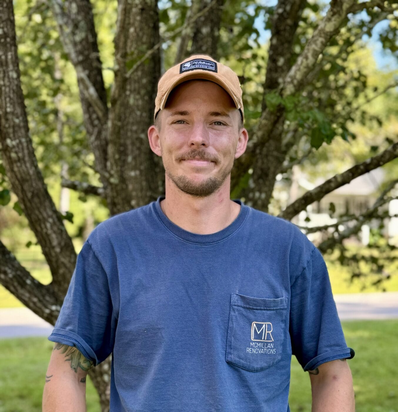 A man in a blue shirt and beige cap stands outdoors in front of a tree, smiling.