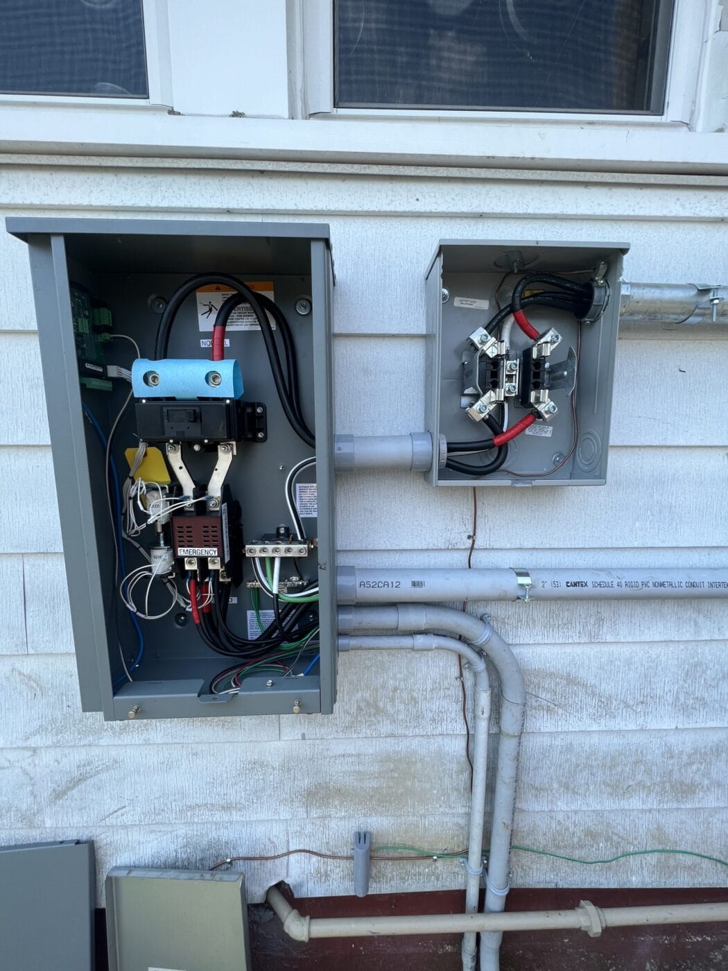 Two open electrical boxes with visible wiring and components are mounted on an exterior wall. Conduit pipes connect to the boxes.