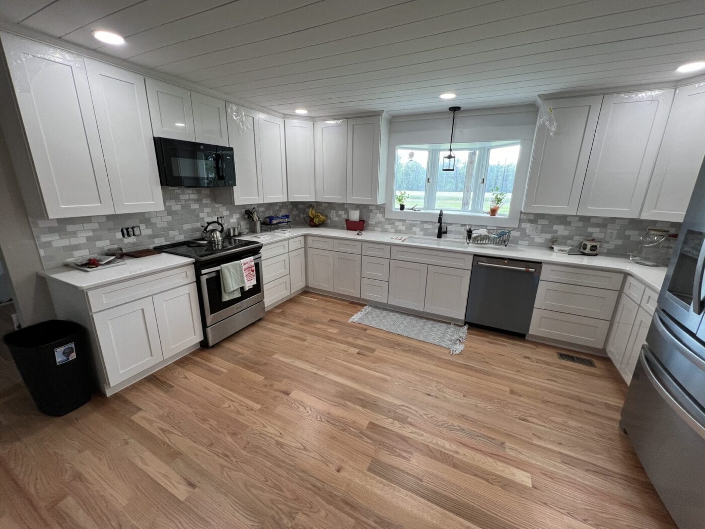 A modern kitchen with white cabinets, stainless steel appliances, hardwood floor, and a window above the sink. A black trash bin is near the corner.