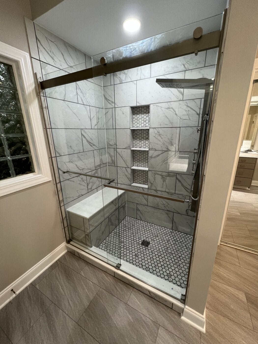 Modern bathroom with a glass-enclosed shower featuring white and gray marble tiles, built-in shelves, a bench, and hexagonal floor tiles.